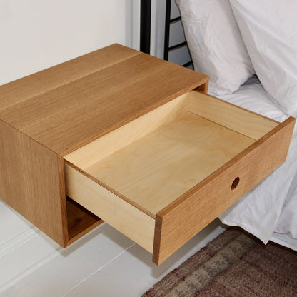 A Krovel Floating Nightstand in White Oak is elegantly mounted beside a bed dressed in white bedding. It includes an open, soft-close drawer with a minimalistic circular pull, and a section of carpet is visible beneath it. 