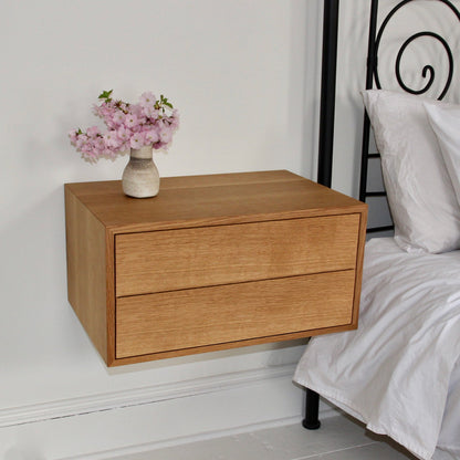 A Krovel Floating Nightstand in White Oak, crafted from solid American White Oak, is mounted beside a bed adorned with white pillows and sheets. It includes two soft-close drawers, with a small vase of pink flowers elegantly resting on top. The room boasts a minimalist design. 