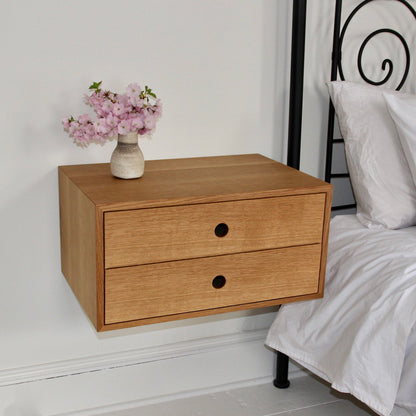 A Floating Nightstand in White Oak by Krovel is mounted on a white wall beside a bed with white linens. Featuring two soft-close drawers, it holds a small vase with pink flowers gracefully atop its surface. 