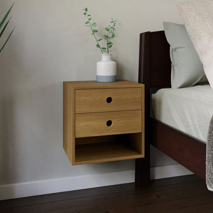 A Krovel Floating Nightstand in White Oak, featuring two soft-close drawers, is mounted beside a bed. It is adorned with a small white vase filled with green foliage on top. The room showcases wooden flooring and a light-colored wall. 