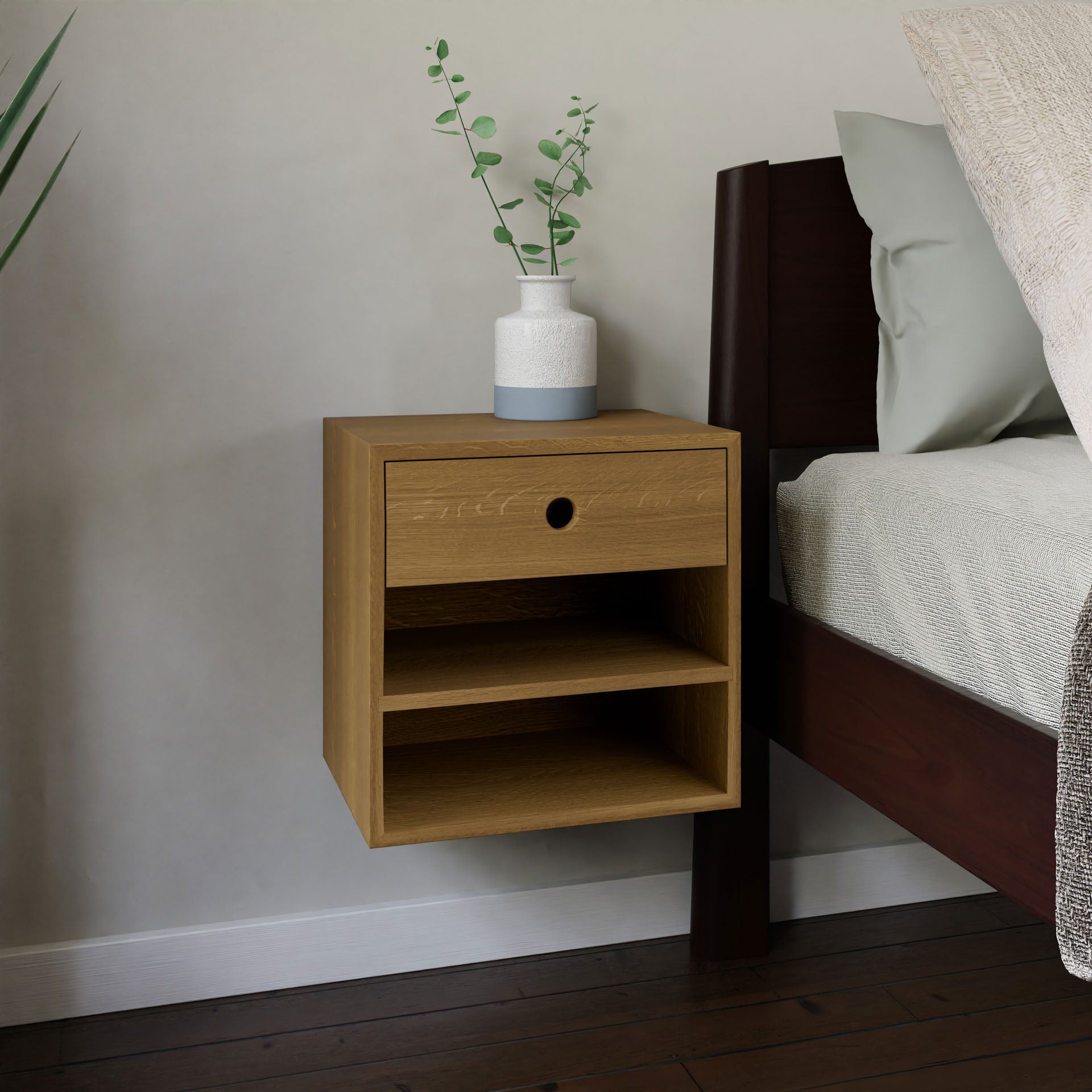 A sleek and contemporary Floating Nightstand in White Oak by Krovel is elegantly mounted on the wall next to a bed. It includes a soft-close drawer and two open shelves. A decorative vase with green foliage adorns the top, while a plant is positioned on the floor nearby. #configuration_single-drawer-double-shelf
