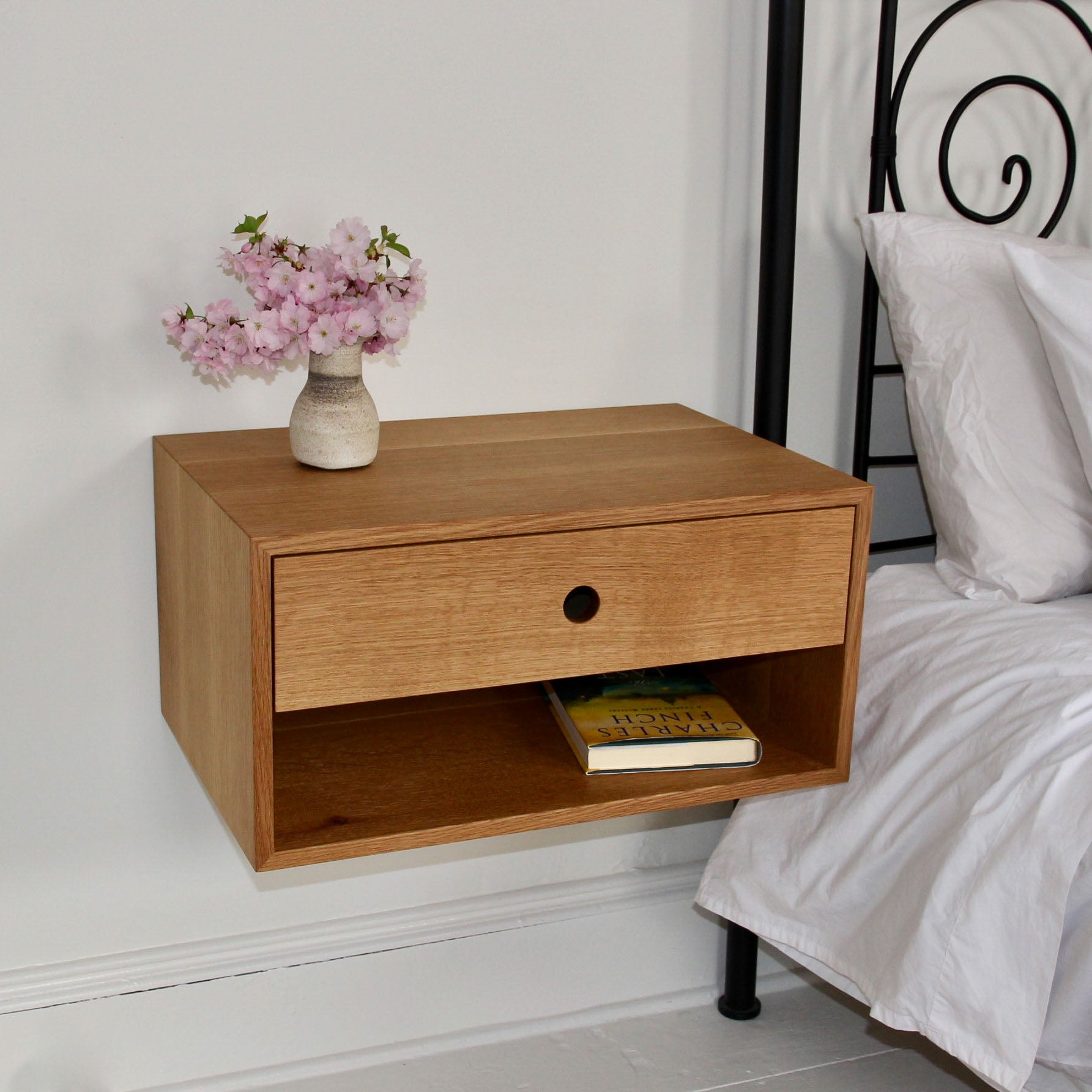 A Krovel Floating Nightstand in White Oak is mounted on a white wall, showcasing its solid American White Oak craftsmanship. It includes a soft-close drawer and an open shelf holding a yellow book. A vase filled with pink flowers decorates the top, while the edge of a bed with white bedding can be seen nearby. #configuration_single-drawer-single-shelf