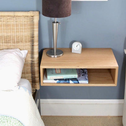A minimalist bedroom showcases a wicker bed headboard and Krovel's Floating Nightstand in White Oak, elegantly crafted from solid American White Oak. A silver lamp is perched on the nightstand, accompanied by books stored in its open shelf. The soft blue walls impart a calming atmosphere, while a small clock elegantly marks time beside the lamp. 