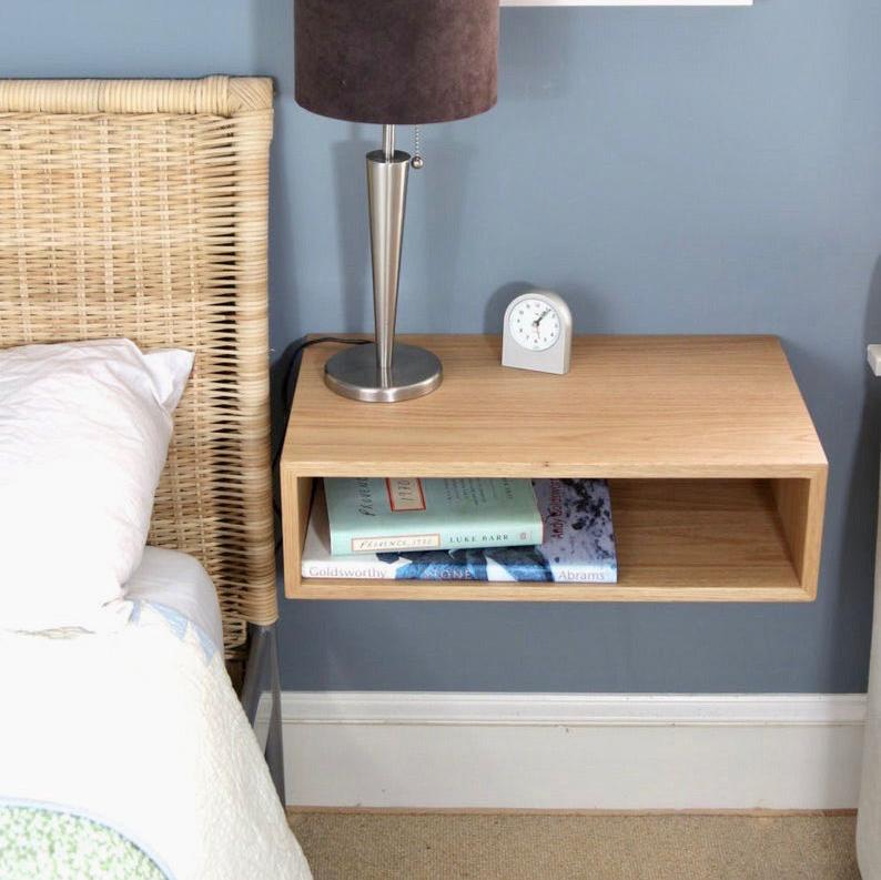 A minimalist bedroom showcases a wicker bed headboard and Krovel's Floating Nightstand in White Oak, elegantly crafted from solid American White Oak. A silver lamp is perched on the nightstand, accompanied by books stored in its open shelf. The soft blue walls impart a calming atmosphere, while a small clock elegantly marks time beside the lamp. #configuration_single-shelf