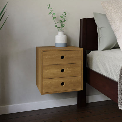 A Krovel Floating Nightstand in White Oak, designed with three soft-close drawers, is mounted beside a dark wood bed. A white and gray vase filled with green foliage rests on top, and the room is completed by a plant in the corner alongside a complementary wooden floor. 