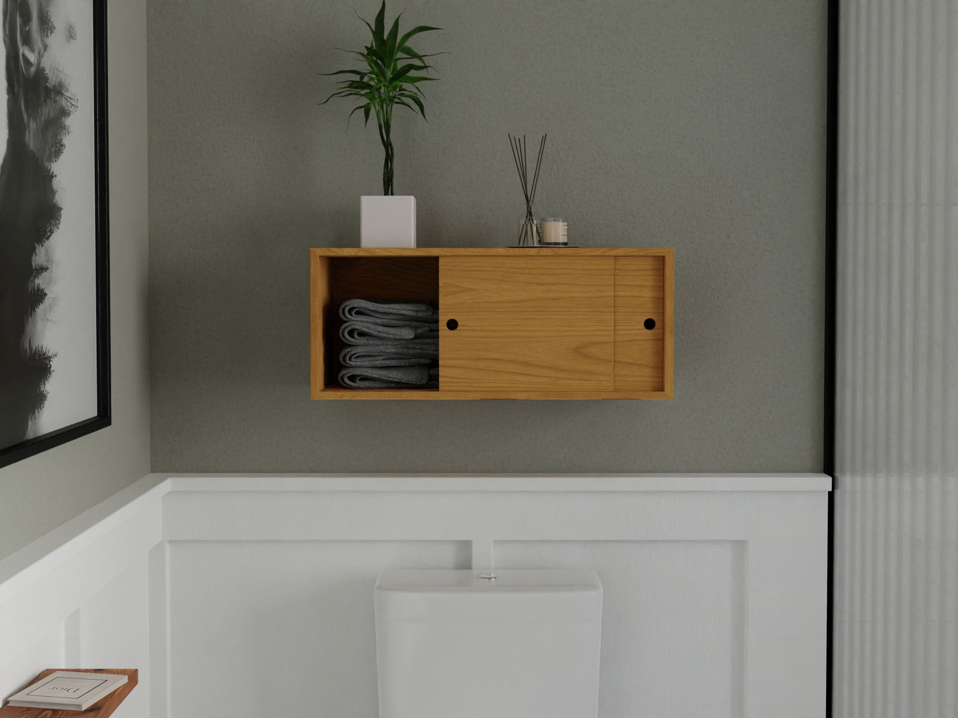 A minimalistic bathroom features a Krovel Floating Storage Cabinet in Cherry, decorated with a small plant, candle, and diffusers. Inside, gray towels are neatly stacked. Below it is a white toilet against a gray and white wainscoted wall with a framed picture partially visible.