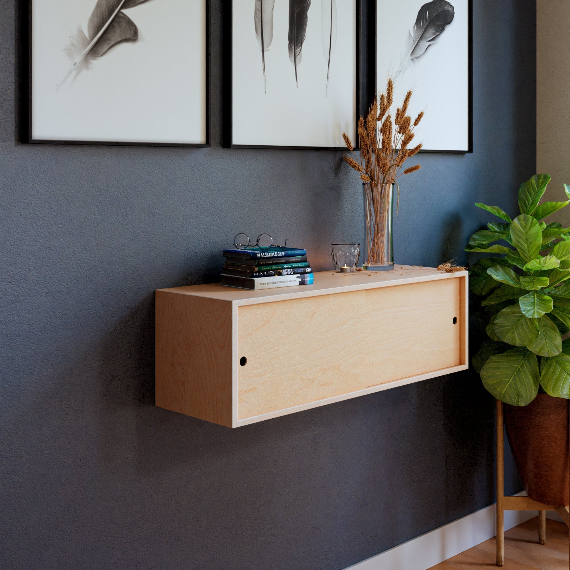 A minimalist room features a Krovel Floating Storage Cabinet in Maple with books, a candle, and a vase of dried flowers. Above it, three framed feather prints are secured by French cleats. A green plant sits on a Northern Hard Maple stand in the corner against a dark wall.