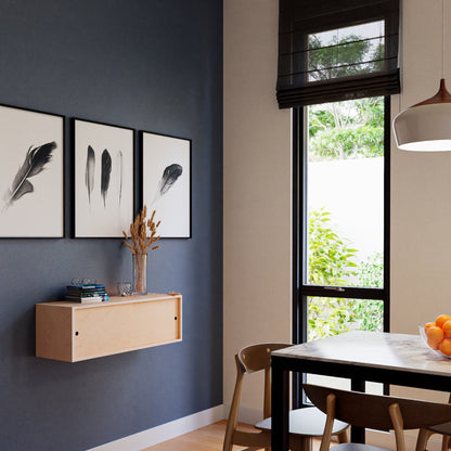 A modern dining area showcases a table with a bowl of oranges, wooden chairs, and a pendant light. Three framed feather prints adorn a gray wall above Krovel's Floating Storage Cabinet in Maple. A large window with black blinds overlooks greenery outside, adding an elegant touch.