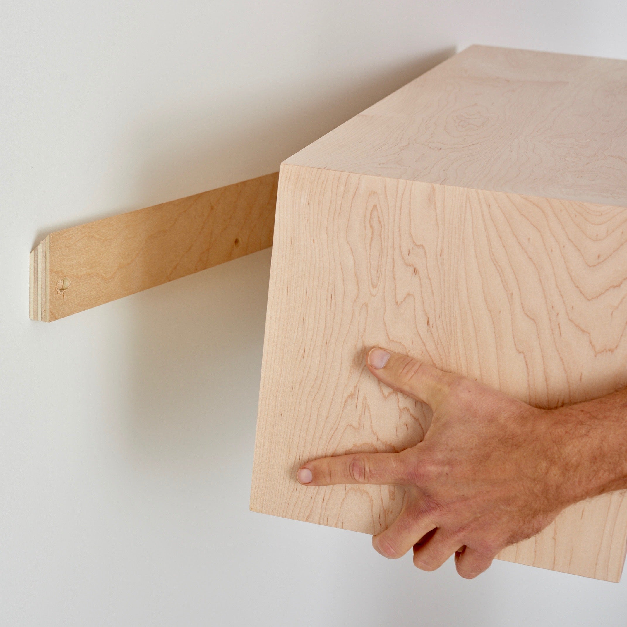 A person is carefully mounting a Krovel Floating Storage Cabinet in Maple onto the wall. Using one hand, they align it to securely fit onto a French cleat bracket, ensuring it's perfectly positioned for a sturdy installation.