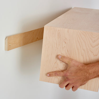 A person is carefully mounting a Krovel Floating Storage Cabinet in Maple onto the wall. Using one hand, they align it to securely fit onto a French cleat bracket, ensuring it's perfectly positioned for a sturdy installation.