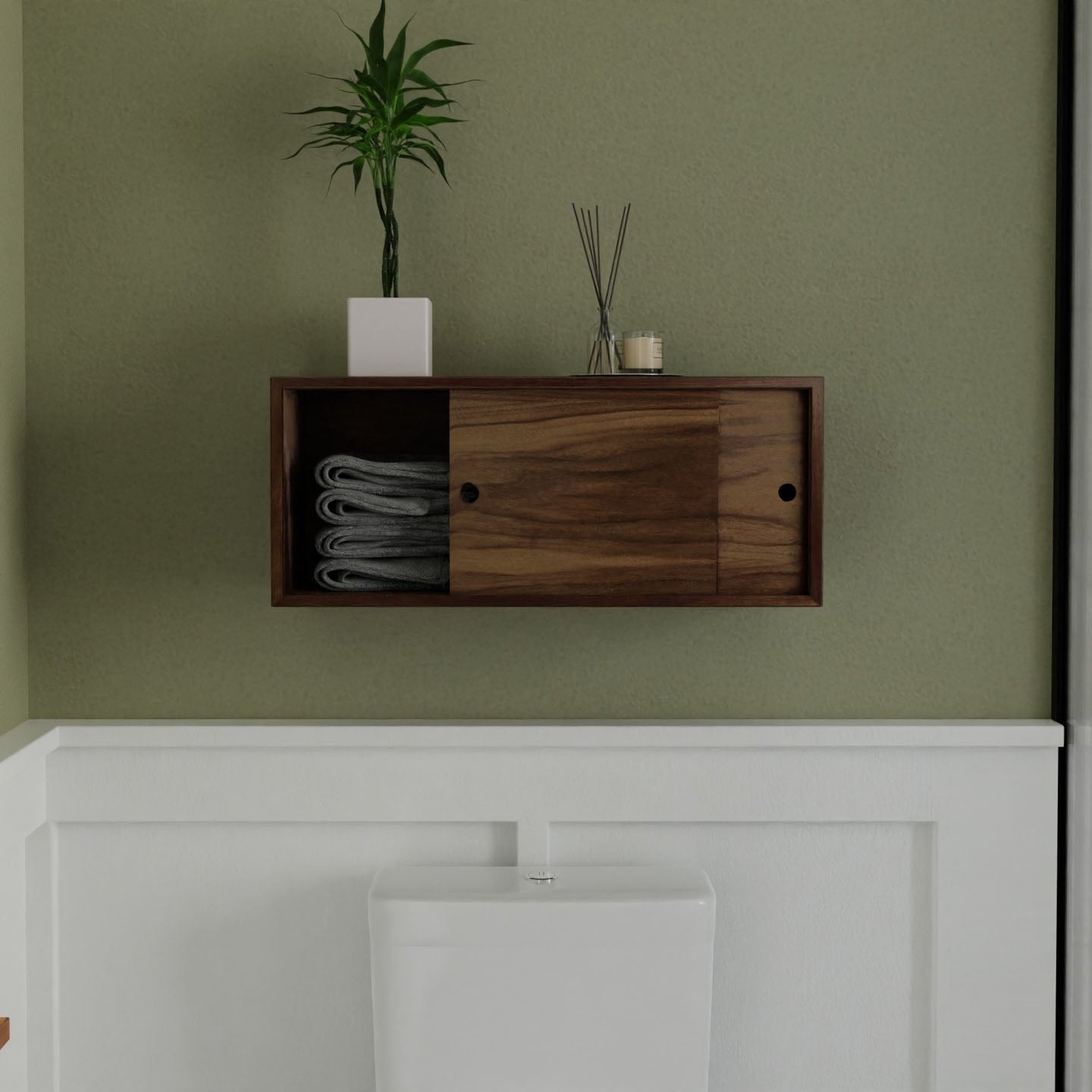 Krovel's Floating Storage Cabinet in Walnut, a handmade small wooden wall cabinet with sliding doors, hovers above a white toilet on a light green wall. It neatly holds gray towels and features a potted plant and small diffuser atop.