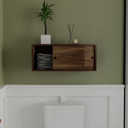 Krovel's Floating Storage Cabinet in Walnut, a handmade small wooden wall cabinet with sliding doors, hovers above a white toilet on a light green wall. It neatly holds gray towels and features a potted plant and small diffuser atop.