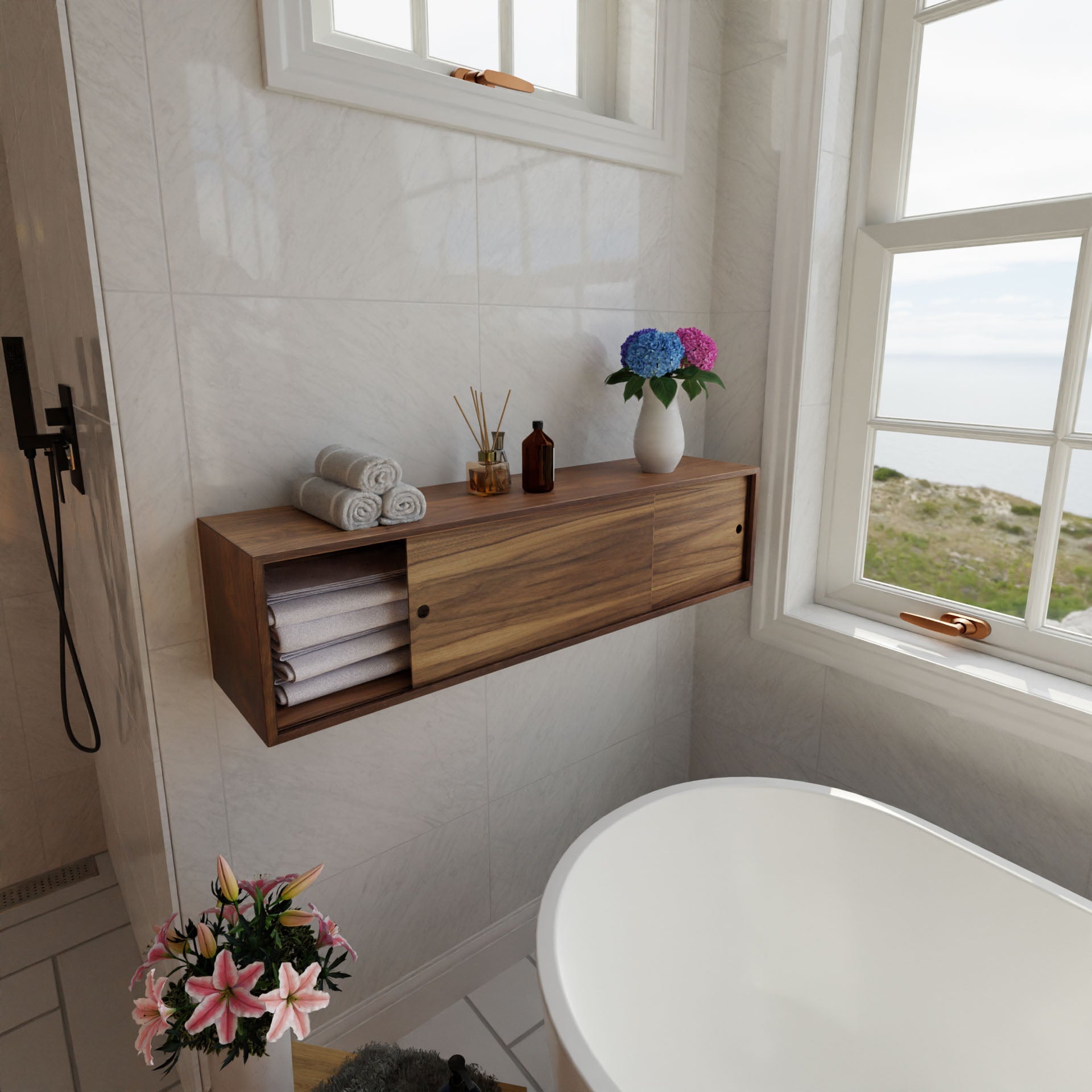 A bathroom showcases a Krovel Floating Storage Cabinet in Walnut, set against a white tiled wall. It stores towels, candles, and a vase with purple and blue flowers. Below an ocean-view window is a white bathtub with a small plant boasting pink flowers nearby.