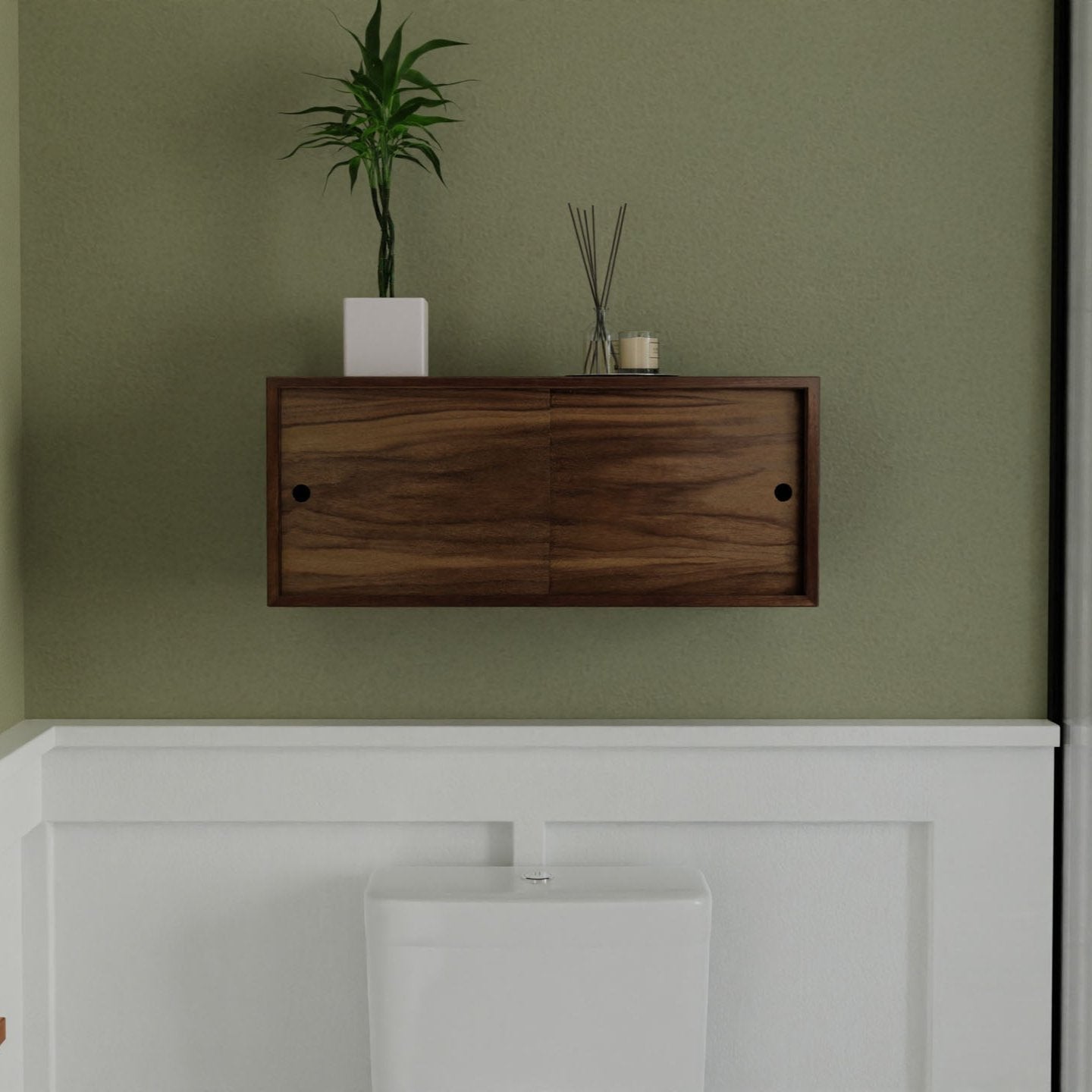 A modern bathroom showcases the Krovel Hardwood Floating Storage Cabinet on the wall, with a green plant in a white pot, reed diffusers, and a candle on top. Below sits a white toilet against light green and white walls.