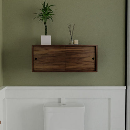 A modern bathroom showcases the Krovel Hardwood Floating Storage Cabinet on the wall, with a green plant in a white pot, reed diffusers, and a candle on top. Below sits a white toilet against light green and white walls.