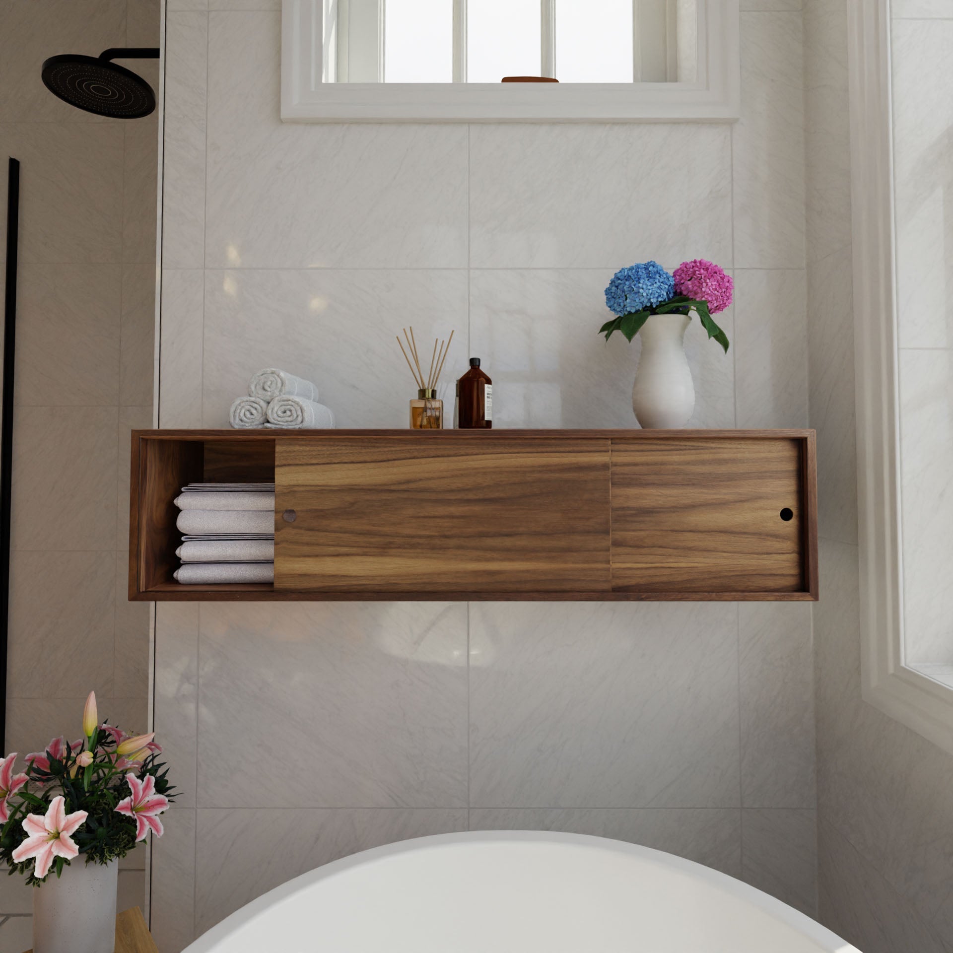 A bathroom with white tiled walls features a Krovel Floating Storage Cabinet in Walnut, holding rolled towels, bottles, a diffuser, and decorated by hydrangeas. Below, the edge of a white bathtub appears. A showerhead and pink lilies are seen in the corner.