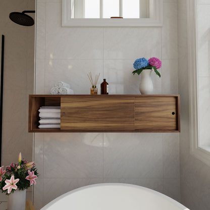 A bathroom with white tiled walls features a Krovel Floating Storage Cabinet in Walnut, holding rolled towels, bottles, a diffuser, and decorated by hydrangeas. Below, the edge of a white bathtub appears. A showerhead and pink lilies are seen in the corner.