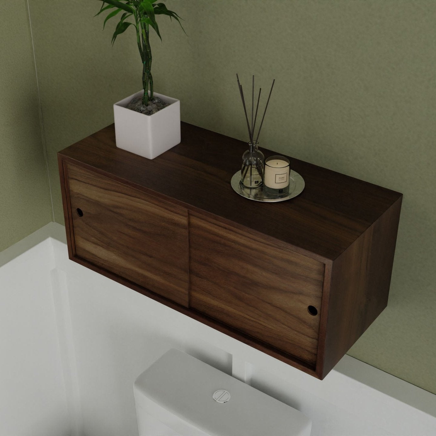 Krovel's Floating Storage Cabinet in Walnut is mounted on a green wall, showcasing its two sliding doors that reveal a small potted plant, reed diffuser, and candle on a metal tray. Below this handcrafted piece is a white toilet with the lid closed.