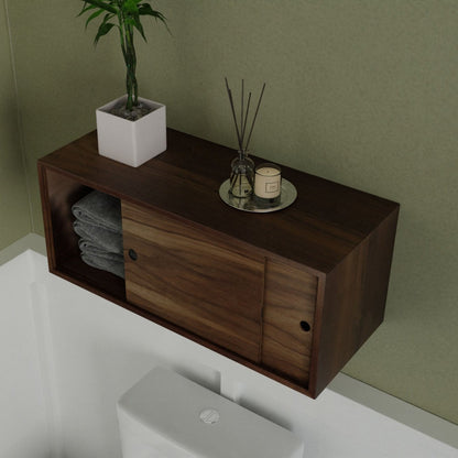 The Krovel Floating Storage Cabinet in Walnut, with sliding doors and towel storage, is handmade from American Black Walnut. Above a toilet against a green wall, it elegantly supports a small potted plant and round-holder scented diffuser on top.