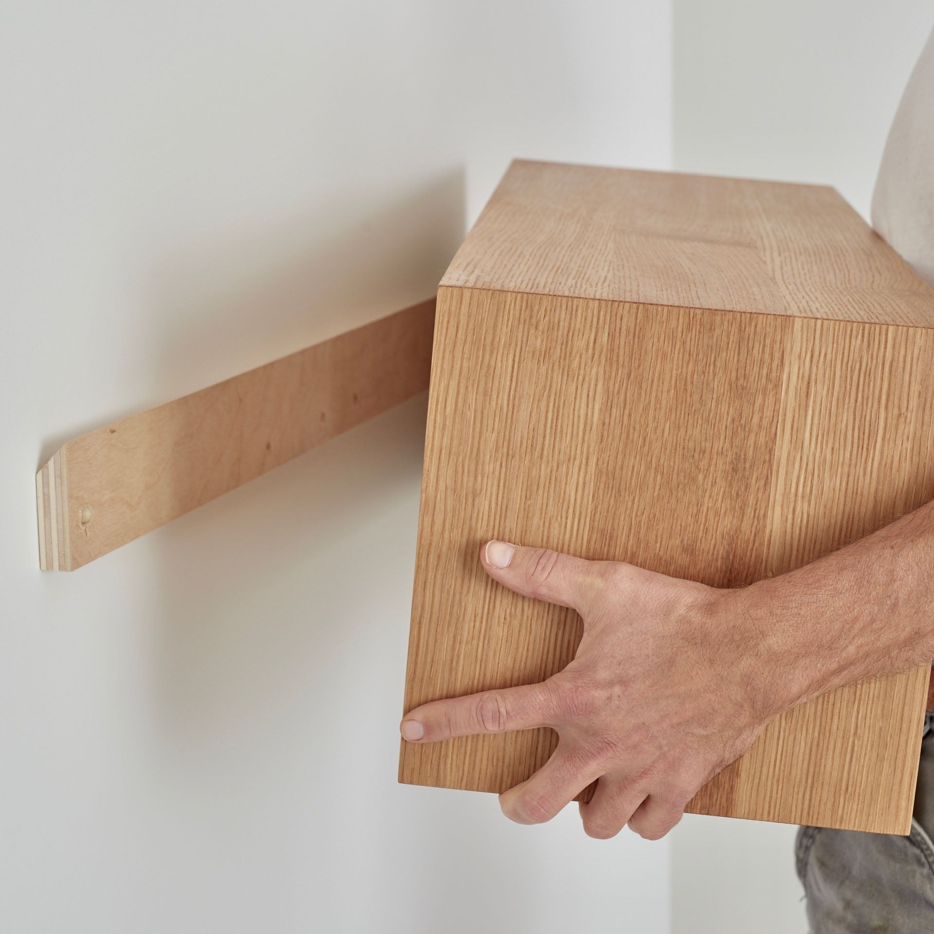 The person expertly mounts the Krovel Floating Storage Cabinet in White Oak using a French cleat, perfectly aligning it with the bracket. Both the wall and cabinet have a light wood grain finish as final adjustments are made.