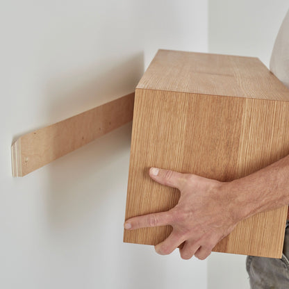 The person expertly mounts the Krovel Floating Storage Cabinet in White Oak using a French cleat, perfectly aligning it with the bracket. Both the wall and cabinet have a light wood grain finish as final adjustments are made.
