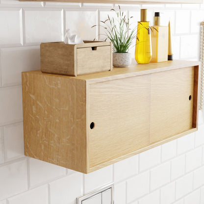 The Krovel Floating Storage Cabinet in White Oak is seamlessly mounted against a white tiled wall, featuring sliding doors that reveal ample space. On top sits a wooden tissue box next to a small plant and vibrant yellow bottles, all crafted for an elegant look.