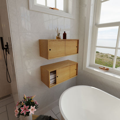 A minimalist bathroom with white walls features elegant wooden shelves and Krovel's Floating Storage Cabinet in White Oak for toiletries. A white bathtub near large windows provides a view of the sea, while a flower arrangement adds a thoughtful touch on the floor.