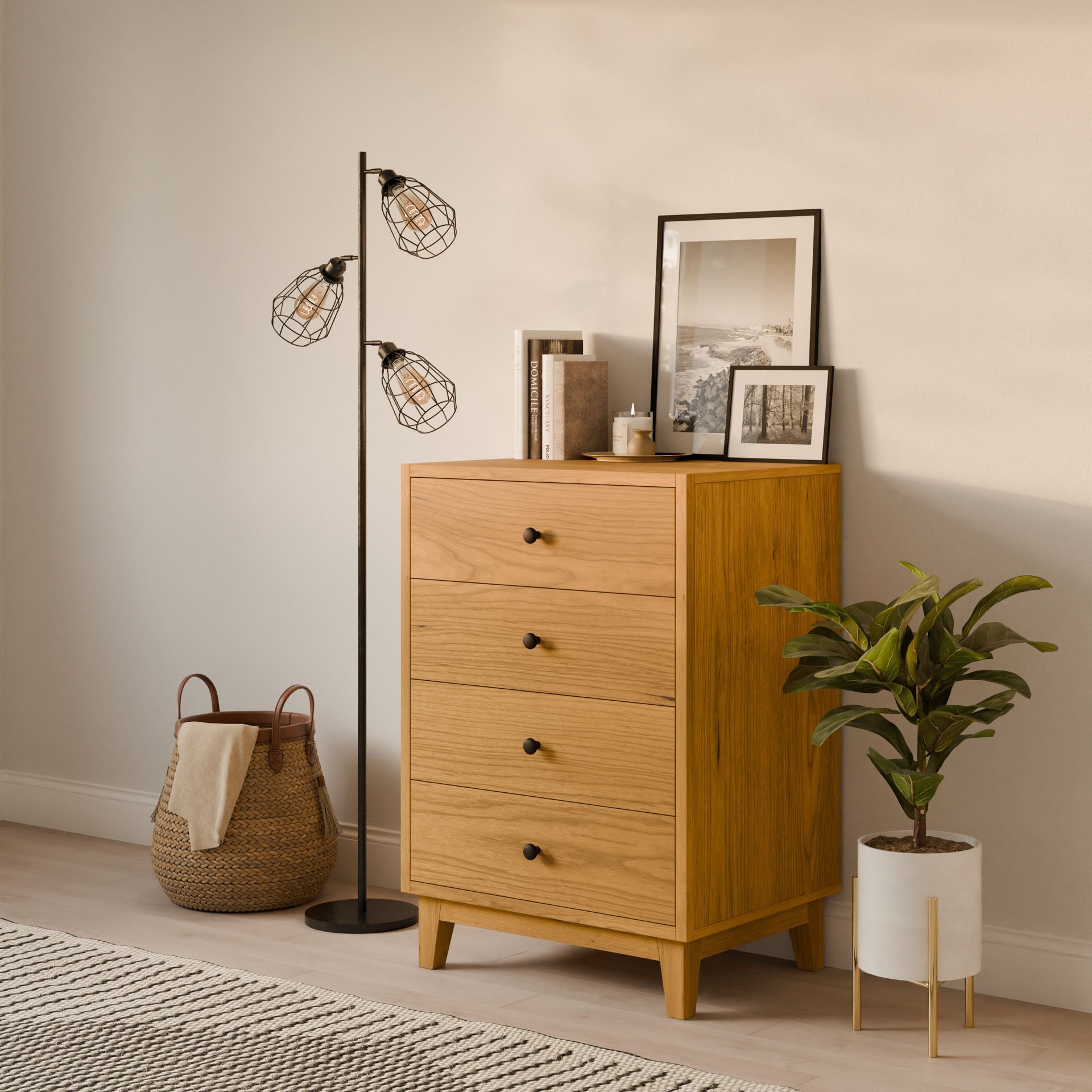 A cozy room corner showcases the Krovel Furniture Co.s Four Drawer Dresser in Cherry, crafted sustainably and adorned with books and framed art. A potted plant is beside it, a wicker basket rests on the floor, and a modern floor lamp with three industrial bulbs stands nearby.