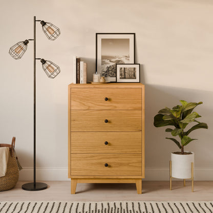 The Krovel Furniture Co. Four Drawer Dresser in Cherry, featuring black knobs, stands beside a potted plant and a black floor lamp with three exposed bulbs. Framed photos and candles adorn this sustainable piece.