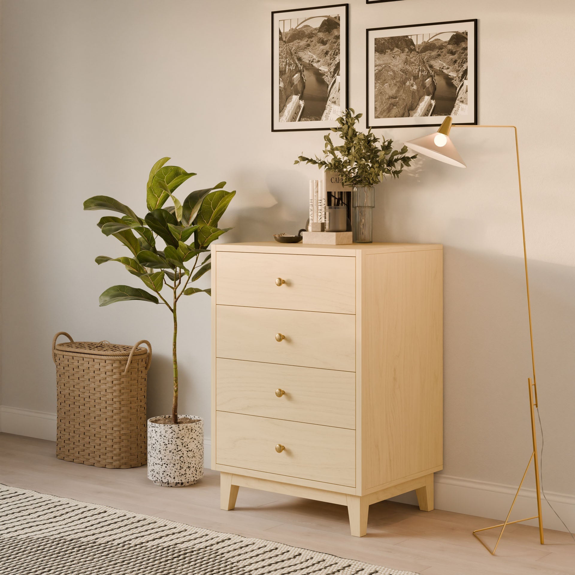 The Four Drawer Dresser in Maple by Krovel Furniture Co., showcasing handmade craftsmanship, stands next to a tall houseplant and wicker basket, illuminated by a gold floor lamp. Two framed photos hang above the dresser, and a small vase with greenery adds charm on top.
