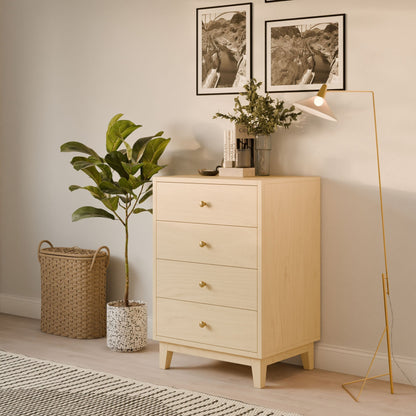The Four Drawer Dresser in Maple by Krovel Furniture Co., showcasing handmade craftsmanship, stands next to a tall houseplant and wicker basket, illuminated by a gold floor lamp. Two framed photos hang above the dresser, and a small vase with greenery adds charm on top.