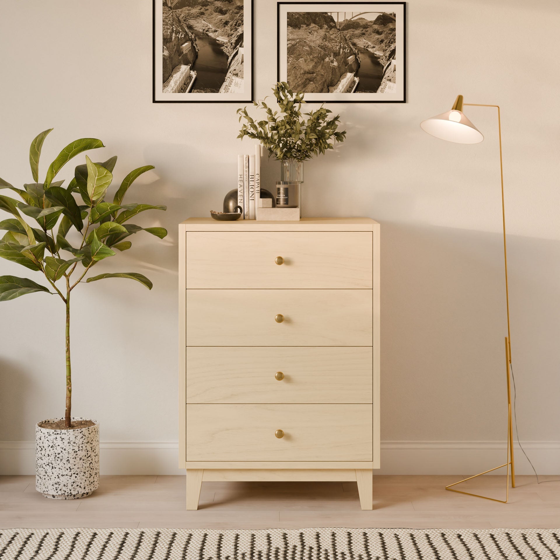 The Krovel Furniture Co. Four Drawer Dresser in Maple stands against a beige wall, displaying its handmade craftsmanship. It is topped with decor and a potted plant. Beside it is a tall floor lamp with a white shade, while another plant sits in a speckled pot below two black-and-white photos.