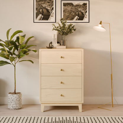 The Krovel Furniture Co. Four Drawer Dresser in Maple stands against a beige wall, displaying its handmade craftsmanship. It is topped with decor and a potted plant. Beside it is a tall floor lamp with a white shade, while another plant sits in a speckled pot below two black-and-white photos.