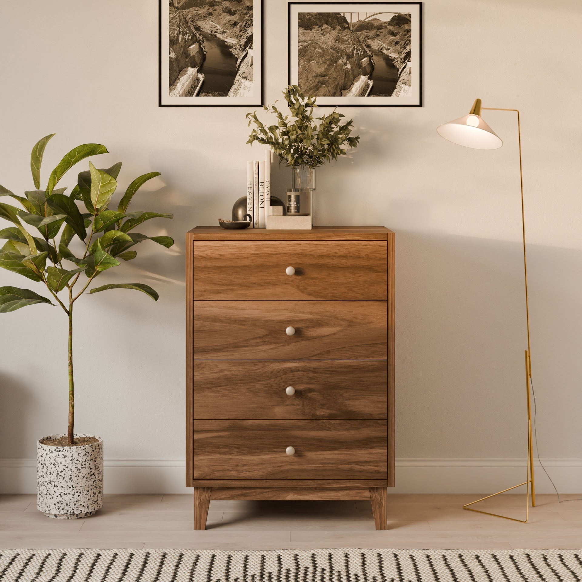 A Krovel Furniture Co. Four Drawer Dresser in Walnut stands against a light wall with two framed photos above it. A gold floor lamp is on the right, a potted plant on the left, and a striped rug enhances its sustainable charm.