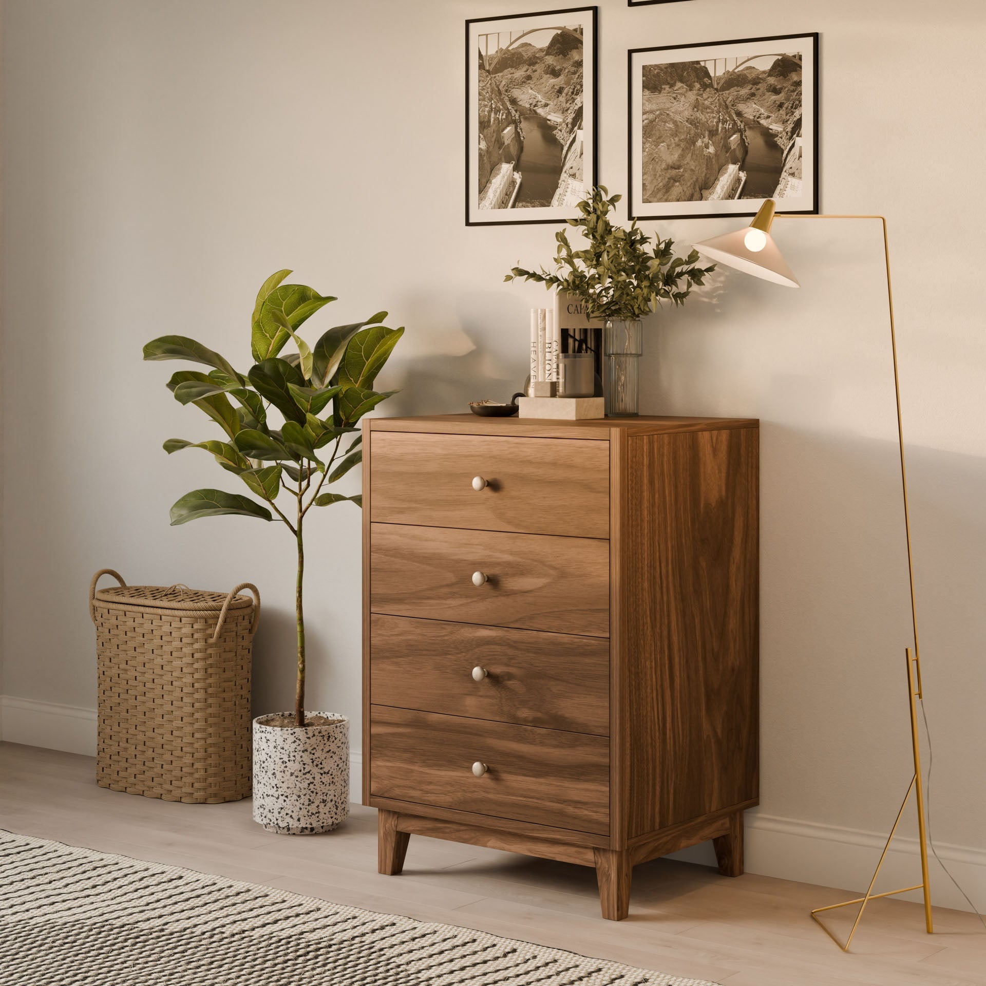The Four Drawer Dresser in Walnut by Krovel Furniture Co. is adorned with books and a potted plant. To the left, a wicker basket with a plant adds charm, while the right features a floor lamp. Above, two framed pictures enhance the rooms minimalist, modern vibe.