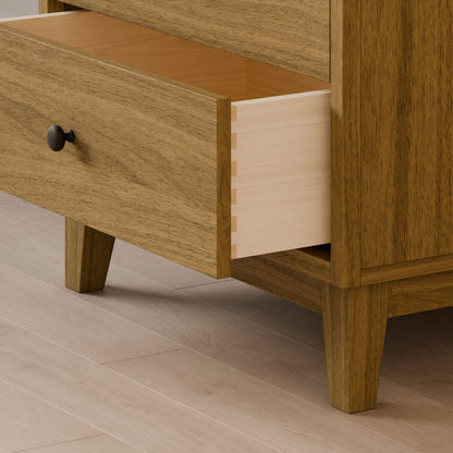 A close-up of the Maine Made Four Drawer Dresser by Krovel Furniture Co. shows dovetail joints on an open drawer. Handmade in Portland, Maine, it stands on four angled legs atop a light wood floor.