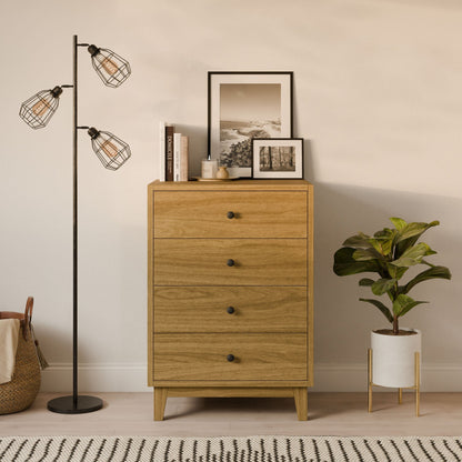 The Four Drawer Dresser in White Oak by Krovel Furniture Co. features sustainable construction and hosts framed pictures and a candle. To the left is a black floor lamp with three exposed bulbs, and to the right is a potted plant on a light rug against a beige wall.