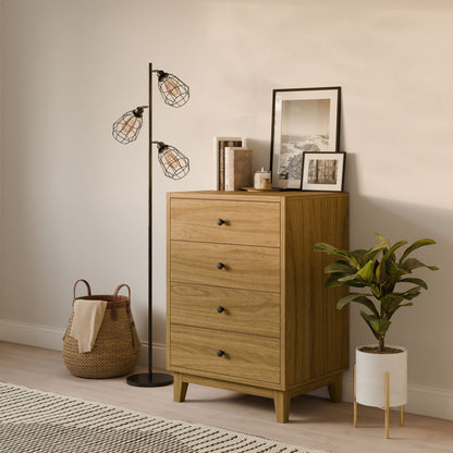 A cozy room corner features a custom-size white oak Four Drawer Dresser by Krovel Furniture Co., a tall black floor lamp with cage bulbs, framed photos, books, a potted plant, and a wicker basket. A textured rug covers the light wood floor.