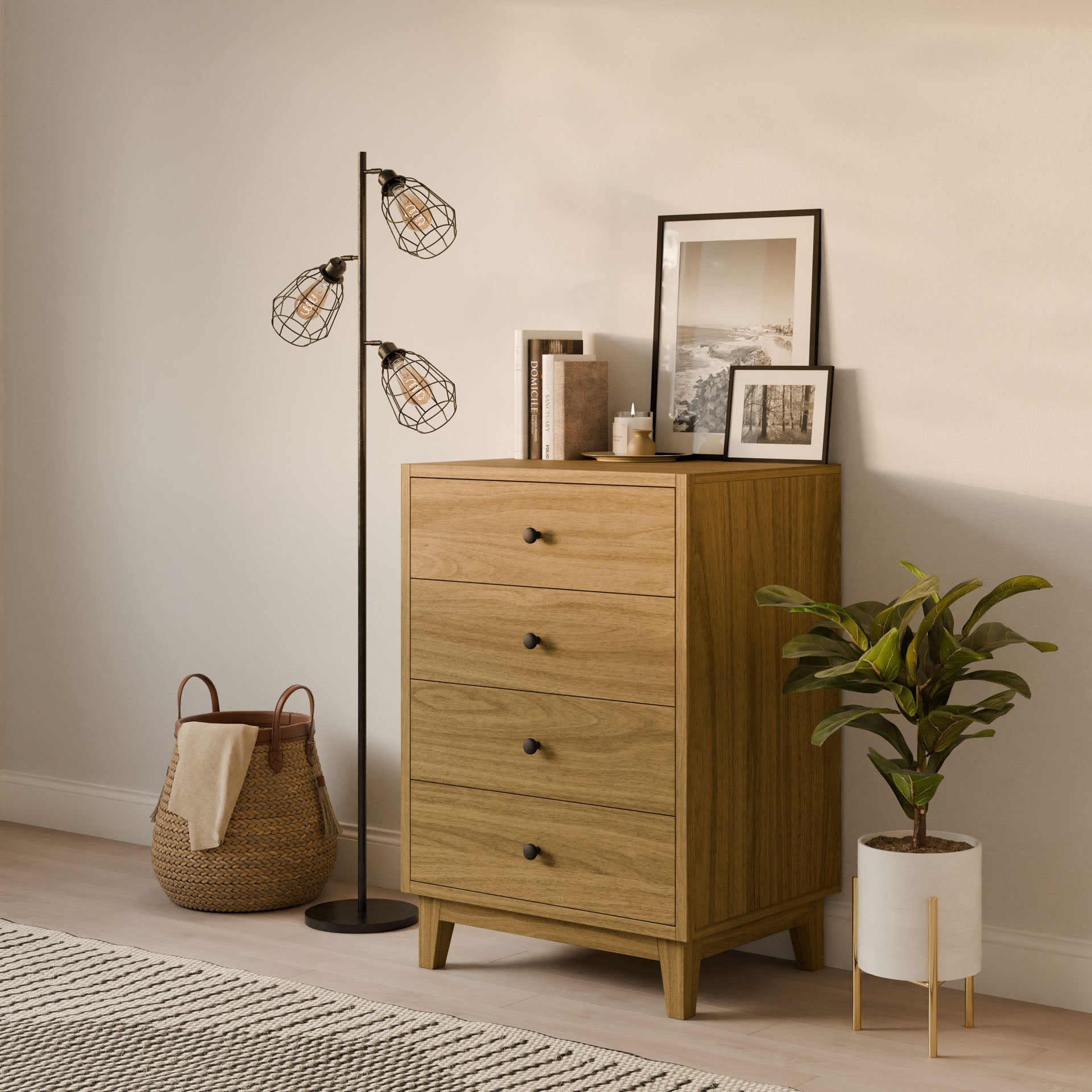 The Krovel Furniture Co. Maine Made Four Drawer Dresser stands beside a black floor lamp with wire cage shades, a potted plant, woven basket with blanket, and framed art and books on top as sunlight streams into the room.