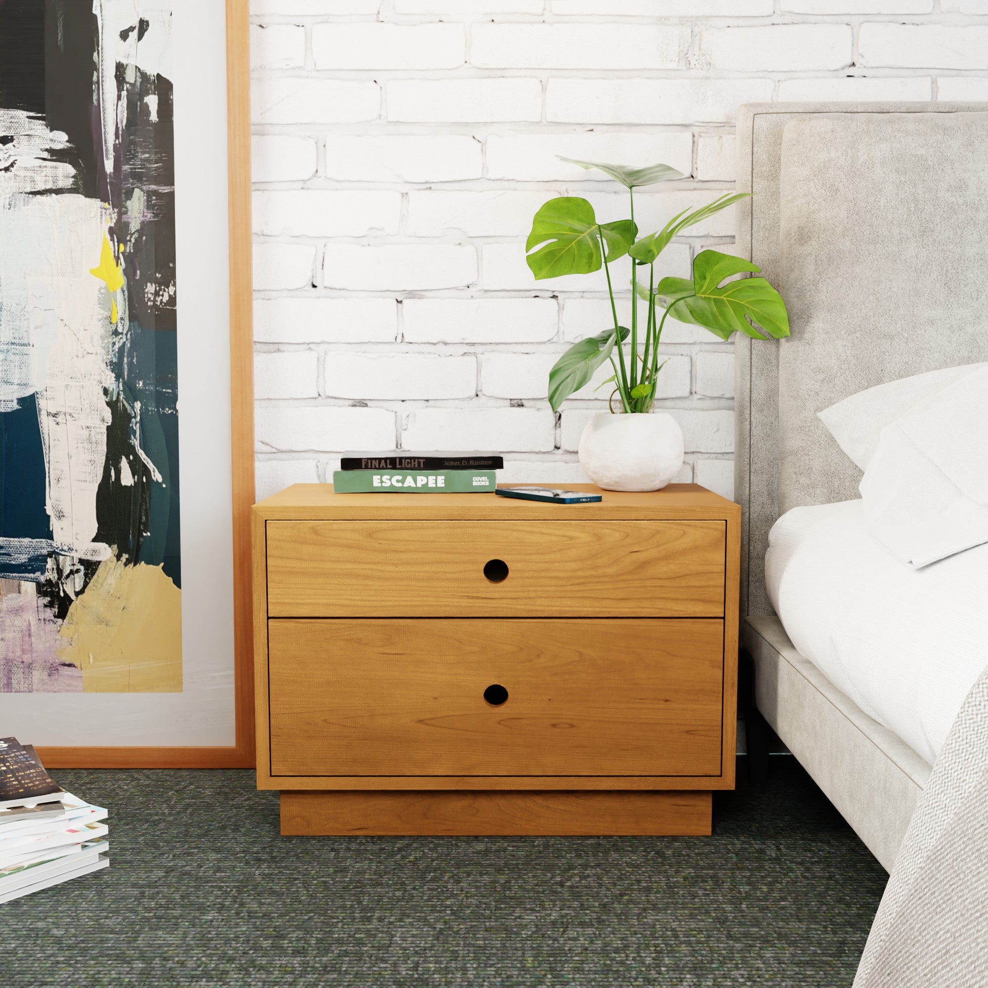 A Krovel Freestanding Nightstand in Cherry, featuring two drawers, stands beside a gray upholstered bed. It holds a potted plant, the book "ESCAPE," and a smartphone. A white brick wall and abstract art set the scene.