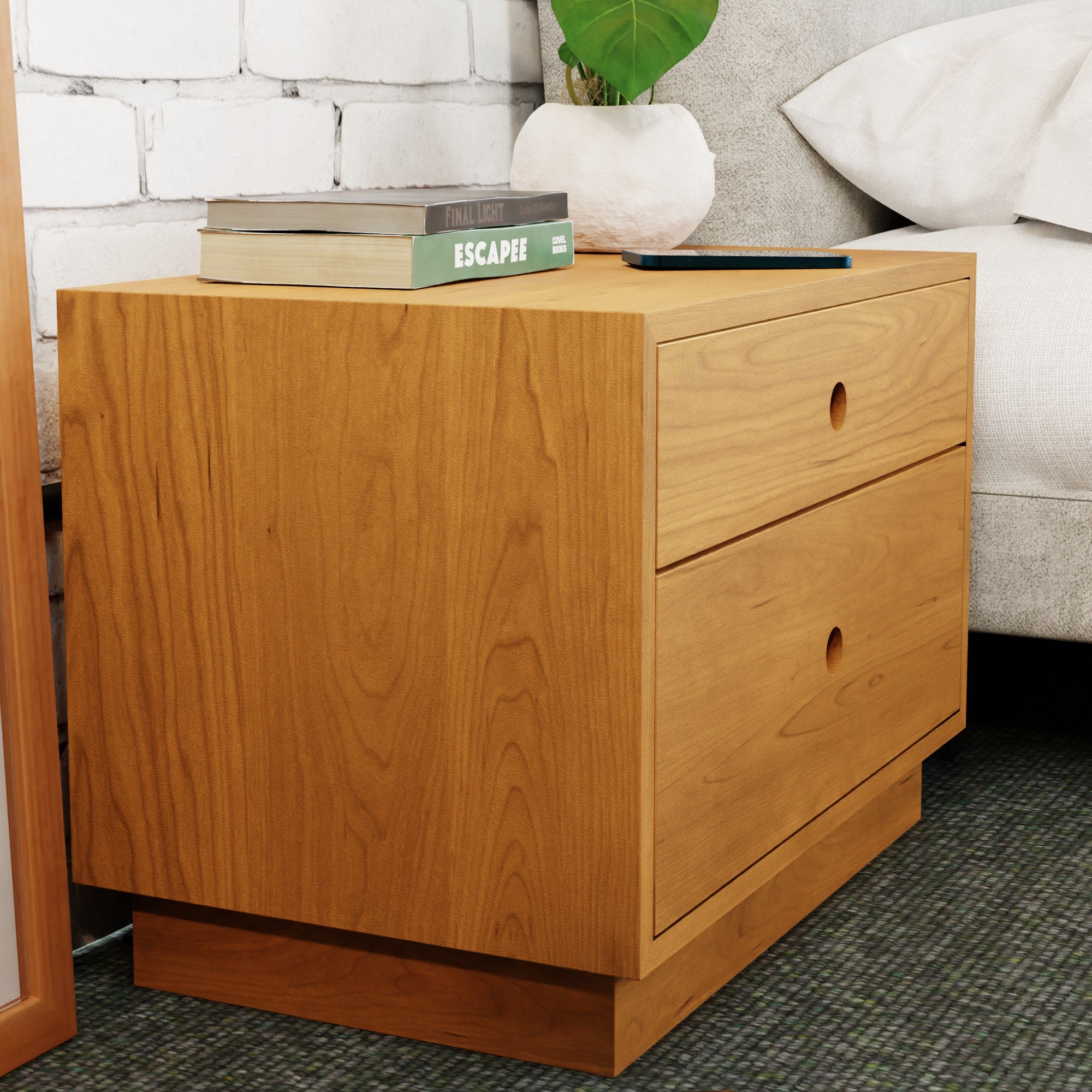 The Krovel Freestanding Nightstand in Cherry, handmade and featuring two drawers, complements the carpeted floor beside the bed. On its surface rest two stacked books and a small plant. The setting is enhanced by a charming white brick wall backdrop.