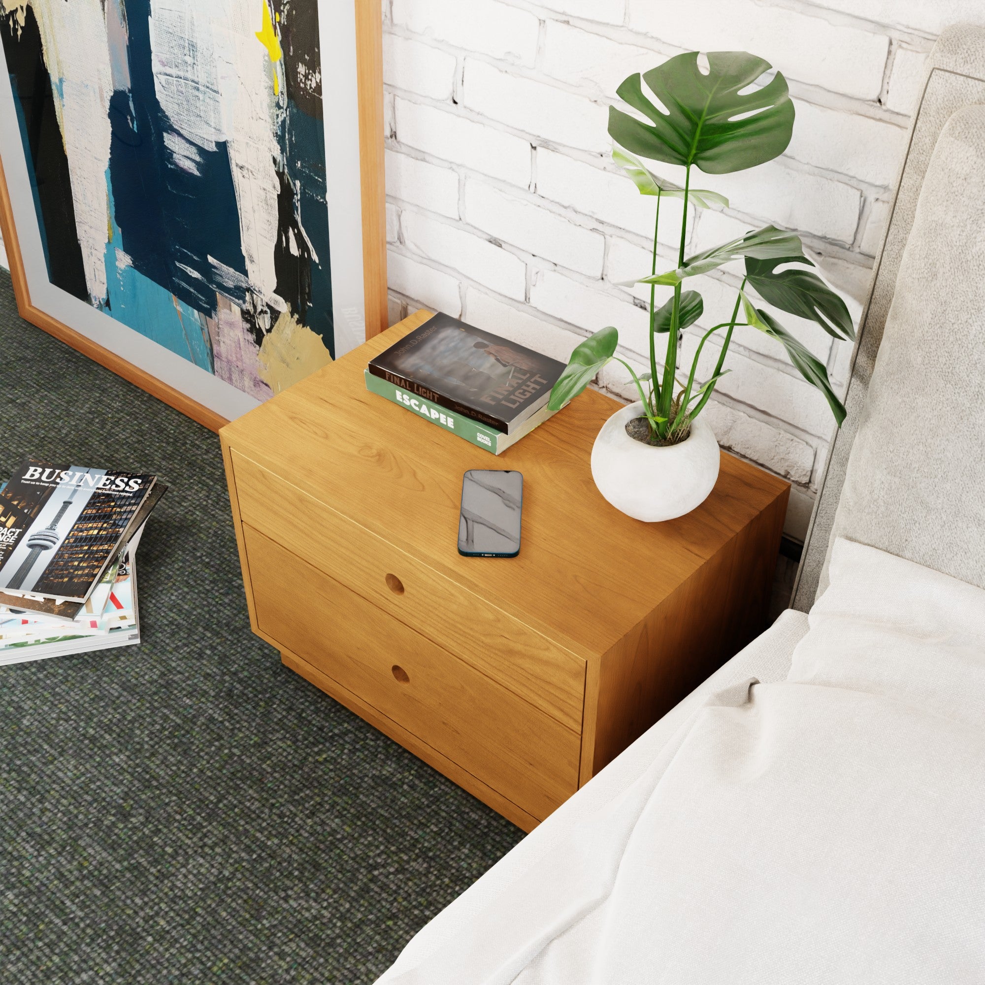 A Krovel Freestanding Nightstand in Cherry with two drawers stands beside the bed, topped with a potted plant, a smartphone, and books titled "Business" and "Escape" beneath an abstract painting propped against the white brick wall, while soft carpeting adds warmth to the room.