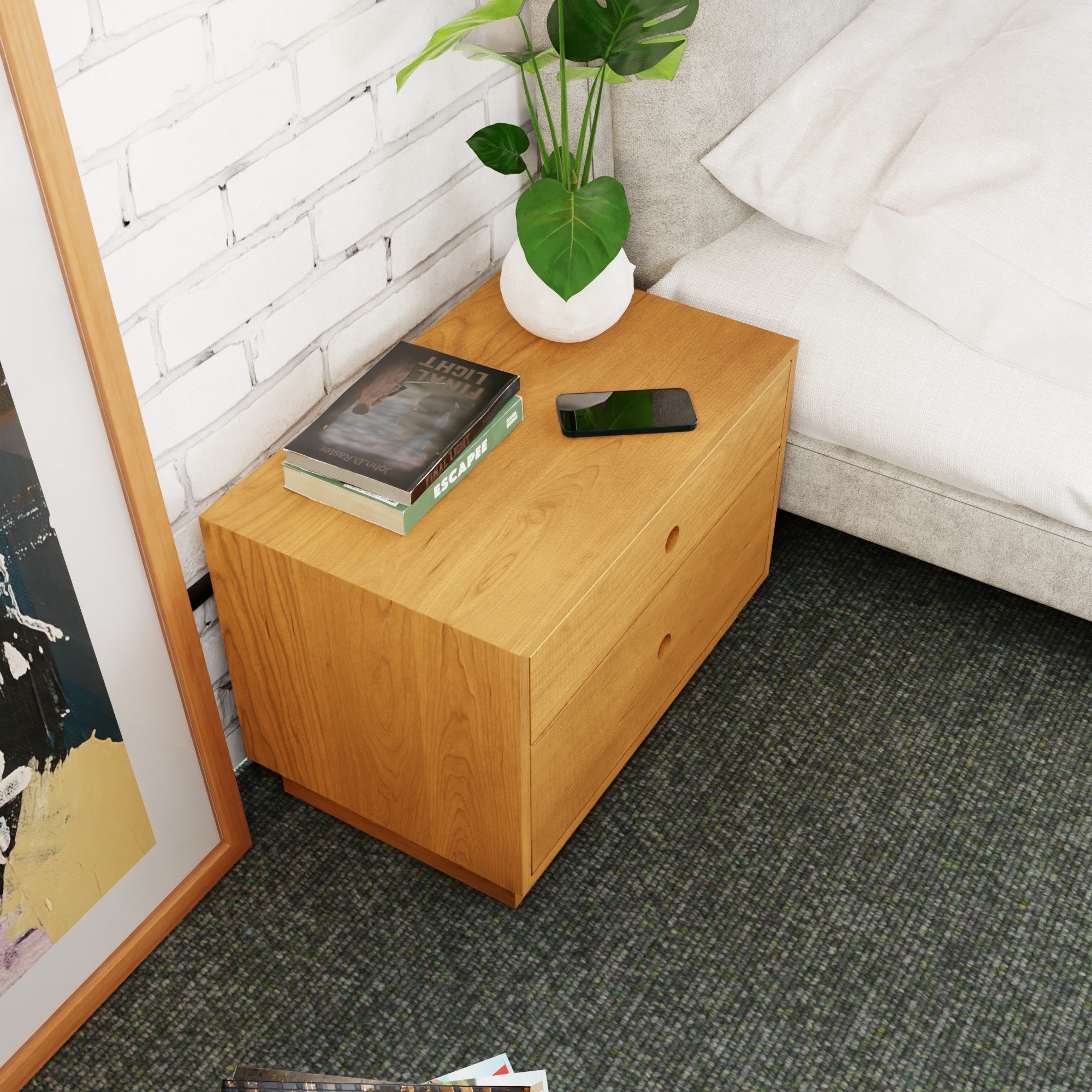 The Krovel Hardwood Freestanding Nightstand with two drawers stands by the bed, topped with a potted plant, two books, and a smartphone. Nearby, part of a framed picture rests against the white brick wall.