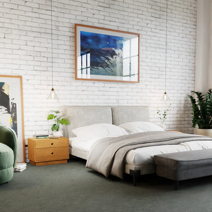 Modern bedroom with a white brick wall, abstract art, light grey upholstered bed, white bedding, Krovel Hardwood Freestanding Nightstand with plants, pendant lights, and a green armchair next to a large leafy plant.