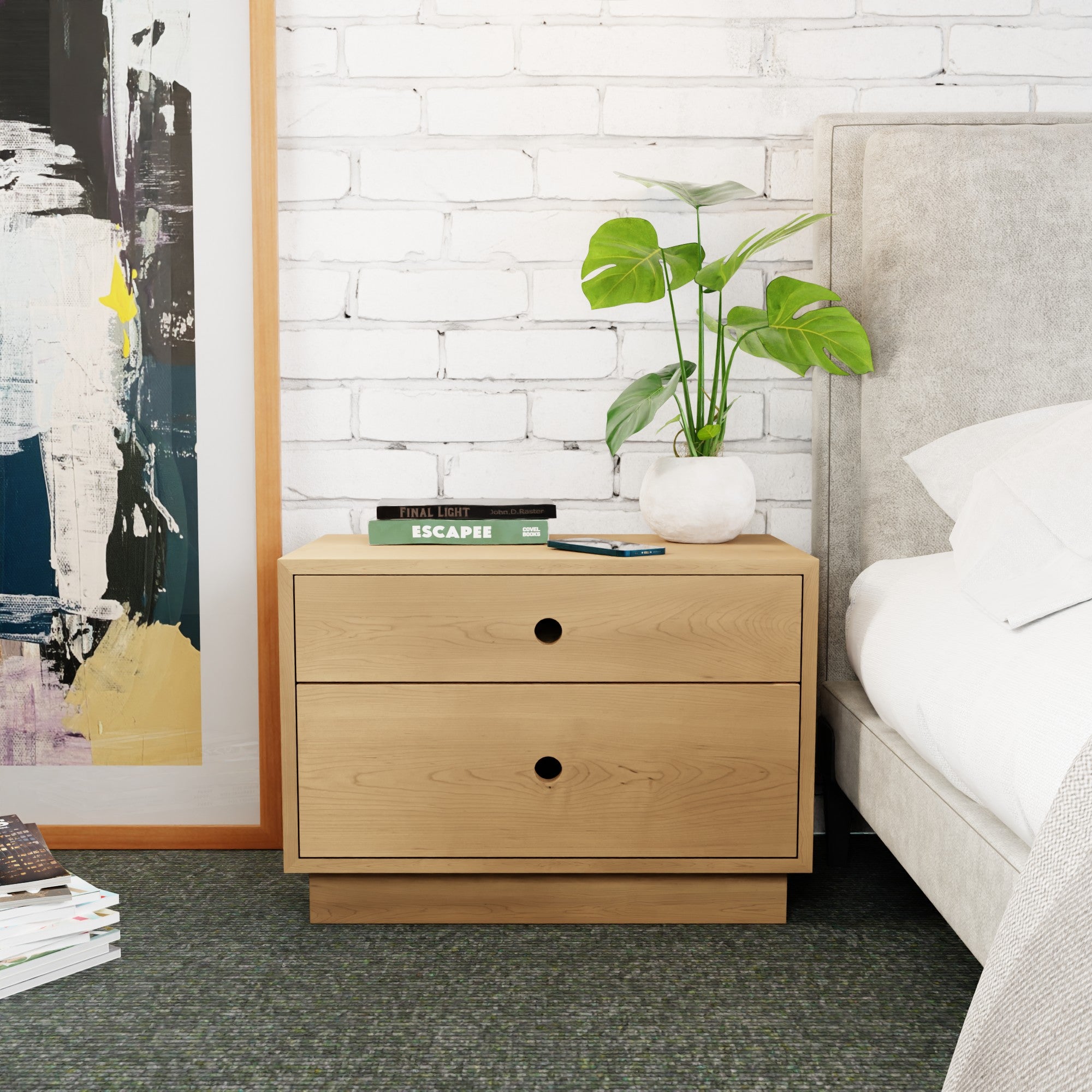 A Krovel Freestanding Nightstand in Maple sits against a white brick wall. It has two soft-close drawers and is adorned with a potted plant, books, and a smartphone. Part of a bed and a colorful abstract painting are visible on the sides.