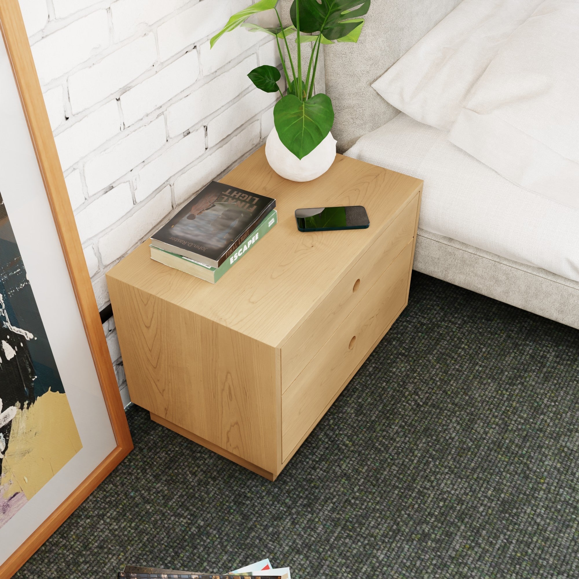A Krovel Freestanding Nightstand in Maple with soft-close drawers sits beside a bed, holding a plant, smartphone, and books. A framed picture rests on the white brick wall behind it, while the dark carpet highlights its elegance.