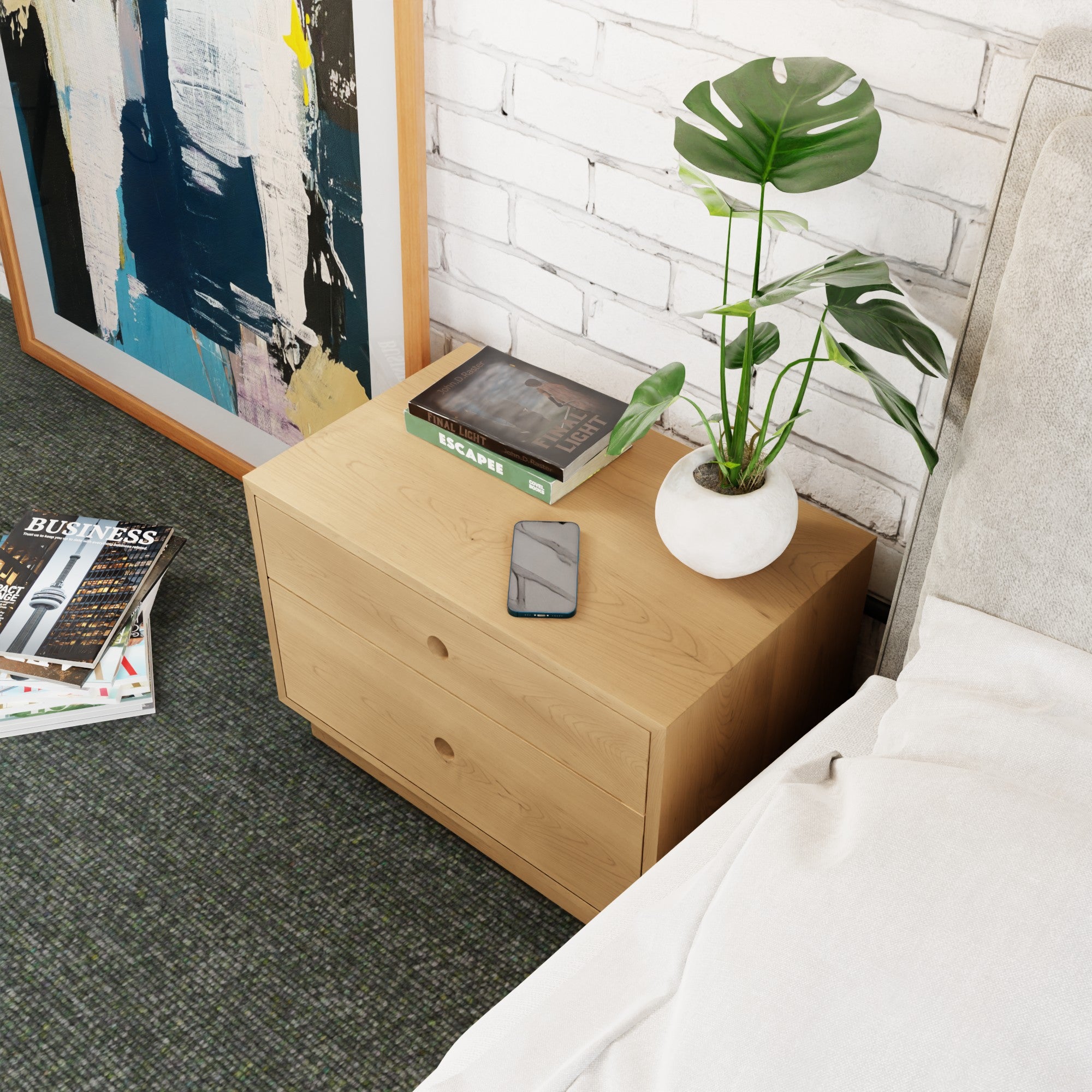A Krovel Freestanding Nightstand in Maple with two soft-close drawers stands beside a bed. A white vase with a potted plant, a smartphone, and the book "Escape" sit on top. Nearby, a painting leans against the white brick wall alongside magazines on the carpeted floor.