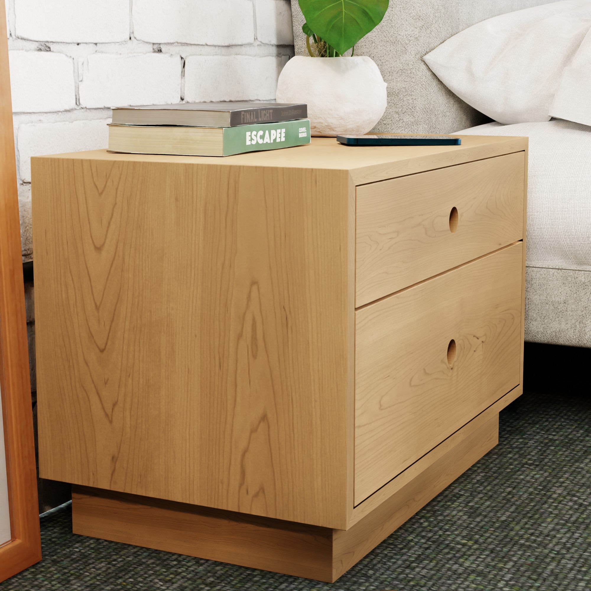 The Krovel Freestanding Nightstand in Maple graces the bedside with its two soft-close drawers for essentials. On top, books rest beside a small green plant, all set against white brick walls and plush carpeted flooring.