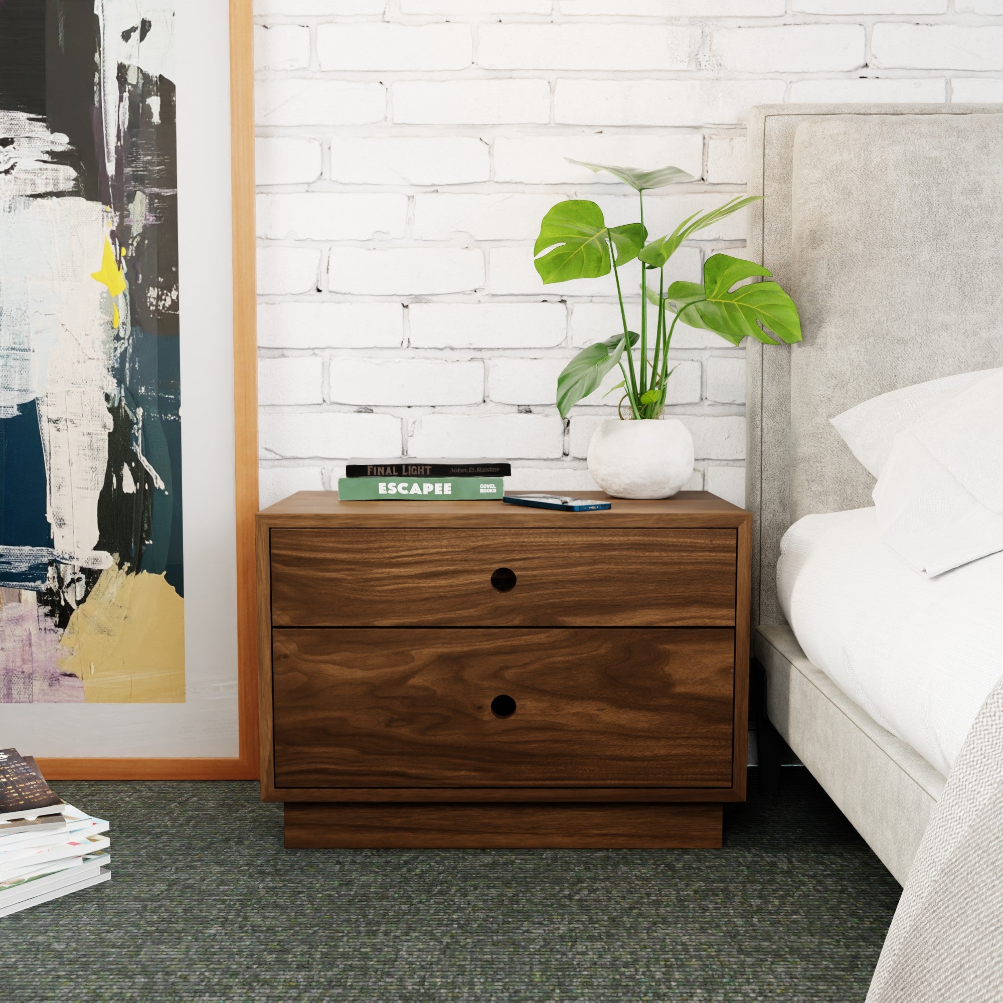 A modern bedroom scene features the Krovel Freestanding Nightstand in walnut, equipped with soft-close drawers next to a beige upholstered bed. It holds a potted plant, phone, and books. A large abstract painting leans on the white brick wall above a soft carpeted floor.