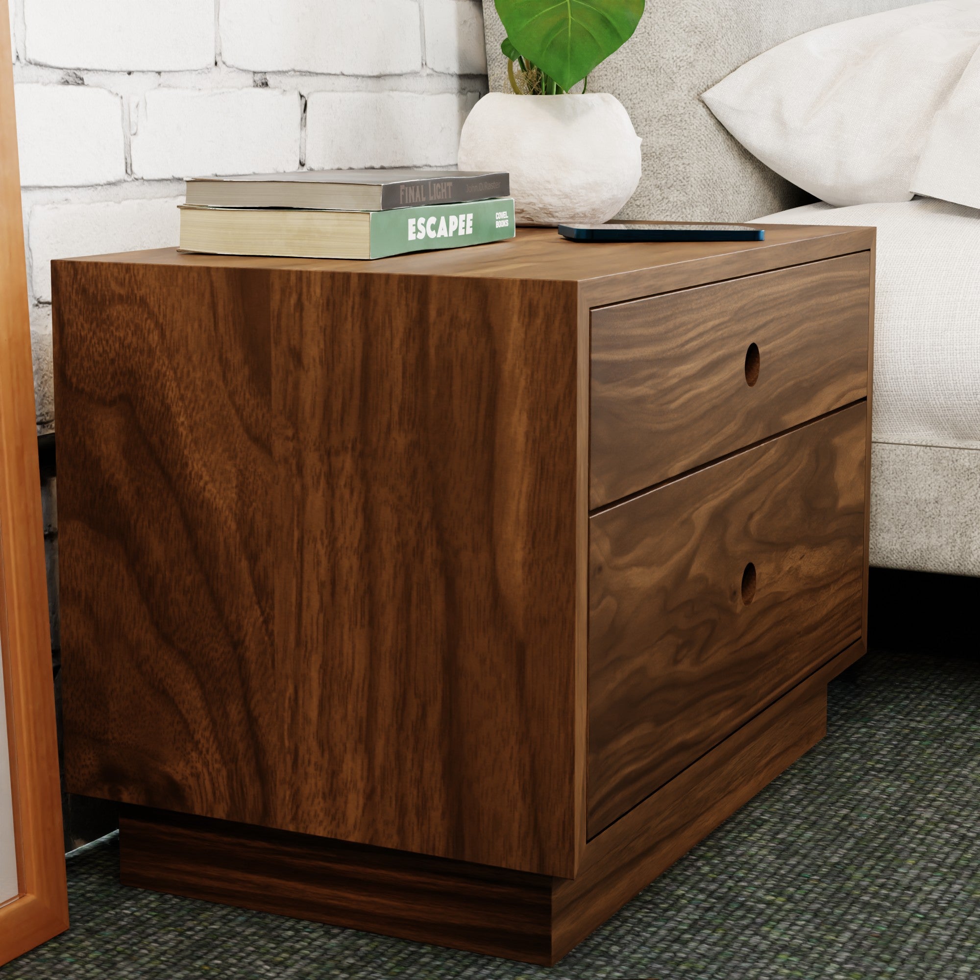 Beside a bed, the Krovel Freestanding Nightstand in Walnut showcases its handmade charm and two soft-close drawers. Books and a tablet adorn its top, set against a white brick wall, with parts of a gray sofa and green plant leaves enhancing the background.